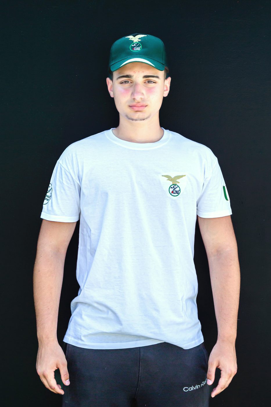 Ragazzo di fronte con maglietta bianca Alpini e cappellino verde